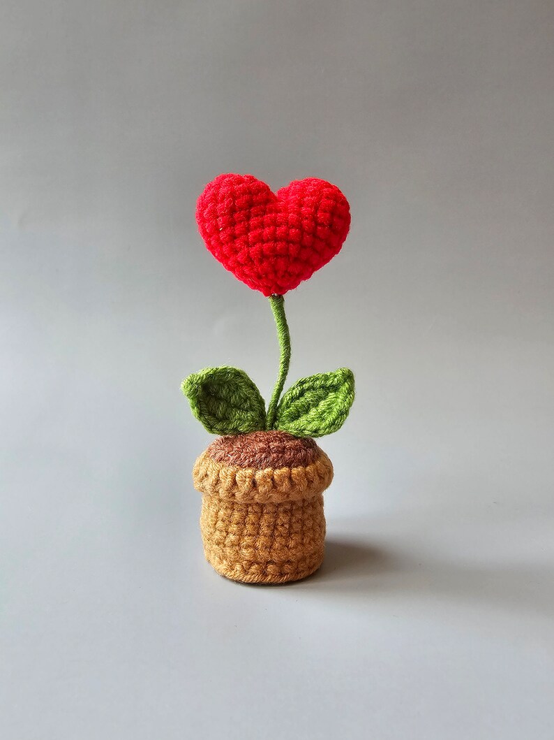 May include: A crocheted heart-shaped flower in a brown pot with green leaves. The flower is red and the pot is brown.
