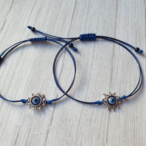 May include: Two blue string bracelets with silver evil eye charms. The charms have a blue eye surrounded by a silver starburst design.