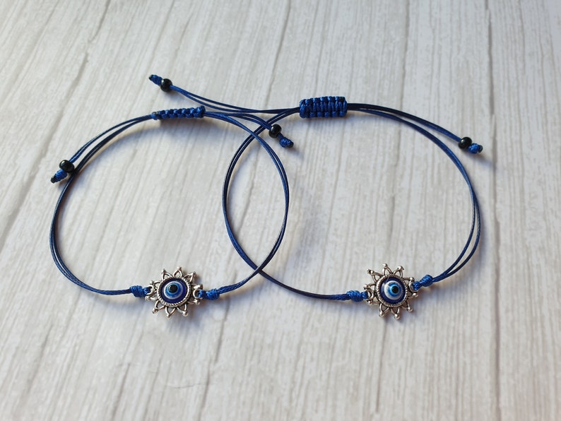 May include: Two blue string bracelets with silver evil eye charms. The charms have a blue eye surrounded by a silver starburst design.