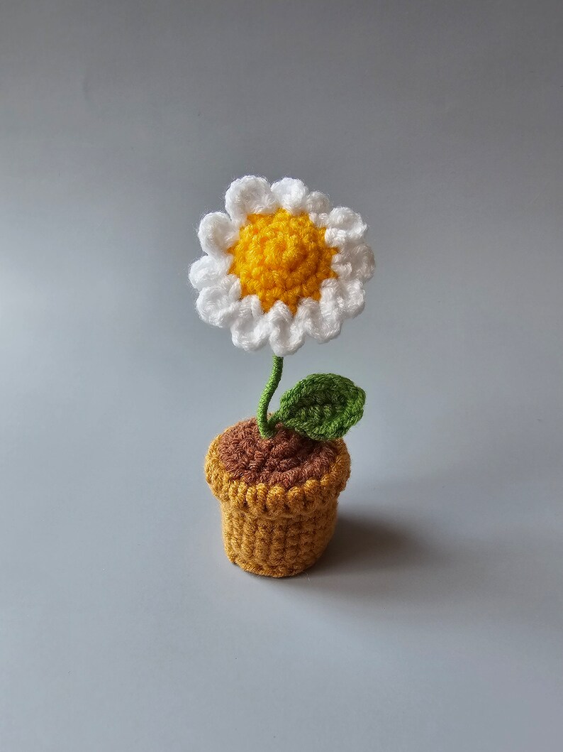 May include: A crocheted daisy flower in a brown and yellow pot. The flower is white with a yellow center and green leaves.