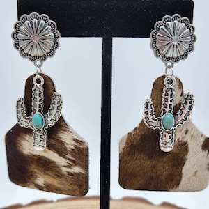 May include: A pair of dangle earrings featuring a brown and white cowhide pattern with a silver cactus charm and a silver concho. The cactus charm has a turquoise stone in the center.