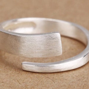 May include: A silver ring with a unique design. The band is a simple circle, but the top is a flat, rectangular piece that wraps around the band.