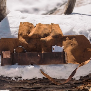 May include: A brown leather roll-up case with a strap, open to reveal a pipe and other small items. The case is lying on a log covered in snow.