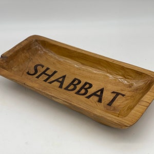 Puede incluir: Una bandeja rectangular de madera para servir con la palabra "Shabbat" grabada en la superficie.