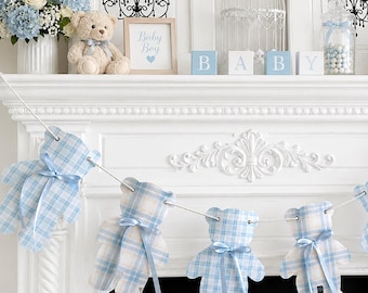 May include: A decorative garland featuring six cutouts shaped like teddy bears, each made from a light blue and white plaid fabric. Every bear cutout is adorned with a small, tied light blue satin ribbon at the neck, all strung together on a white cord.