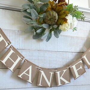 K&ouml;nnte beinhalten: Ein braunes Papierbanner mit dem Wort "THANKFUL" in wei&szlig;en Buchstaben. Das Banner ist mit einer Schnur befestigt und mit einem Herbstkranz verziert.