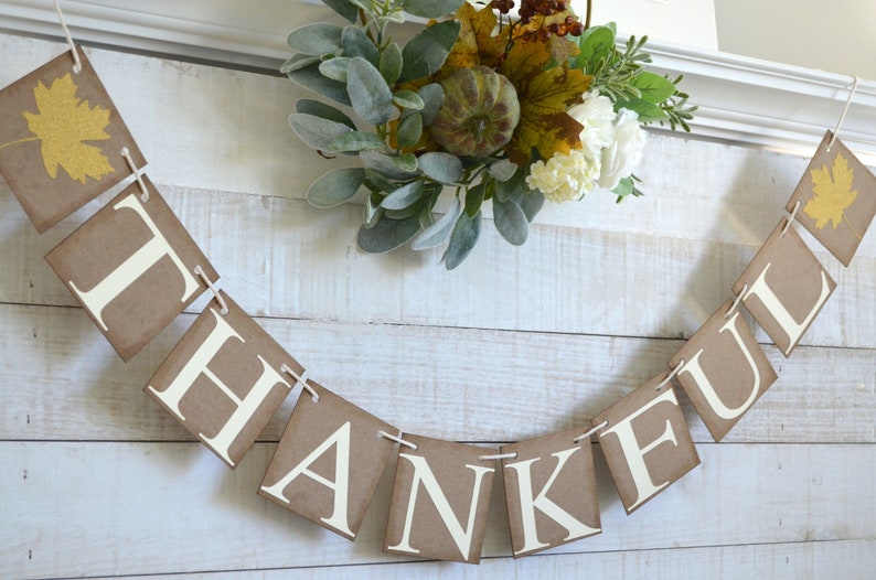 K&ouml;nnte beinhalten: Ein braunes Papierbanner mit dem Wort "THANKFUL" in wei&szlig;en Buchstaben. Das Banner ist mit einer Schnur befestigt und mit einem Herbstkranz verziert.