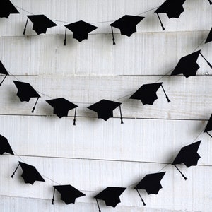 May include: Black graduation cap cutouts strung together on a white string to create a festive garland. The garland is hanging on a white wood background.