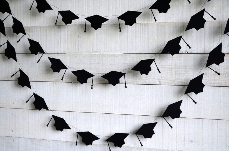 May include: Black graduation cap cutouts strung together on a white string to create a festive garland. The garland is hanging on a white wood background.
