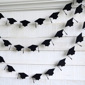 May include: Black graduation cap paper cutouts strung together to form a garland. The garland is hanging on a white wood wall.