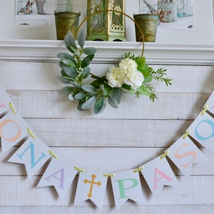 May include: A white wooden mantel with a banner that reads "BUONA PASQUA" in pastel colors. The banner is strung with yellow ribbon. A gold metal wreath with white flowers and green leaves is above the banner.