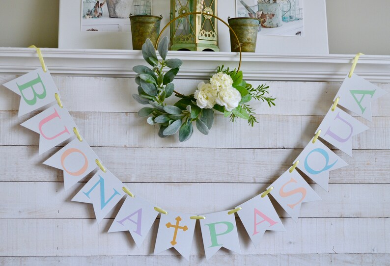 May include: A white wooden mantel with a banner that reads "BUONA PASQUA" in pastel colors. The banner is strung with yellow ribbon. A gold metal wreath with white flowers and green leaves is above the banner.