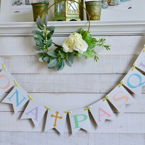 May include: A white wooden mantel with a banner that reads "Buona Pasqua" in pastel colors. The banner is strung with yellow ribbon and has a gold cross in the center. A green wreath with white flowers hangs above the banner.