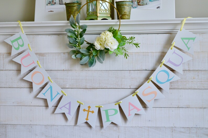 May include: A white wooden mantel with a banner that reads "Buona Pasqua" in pastel colors. The banner is strung with yellow ribbon and has a gold cross in the center. A green wreath with white flowers hangs above the banner.