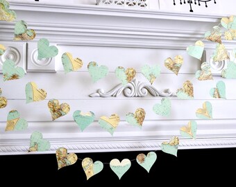 May include: A garland of heart-shaped decorations. Each heart is cut from paper printed with a vintage map design in shades of blue, green, and tan. The garland is strung across a white mantelpiece.