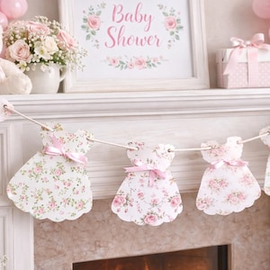 May include: A baby shower decoration featuring a garland of dress-shaped cutouts with pink floral patterns and pink bows. A framed sign reads "Baby Shower" above a fireplace, with pink balloons and gifts in the background.
