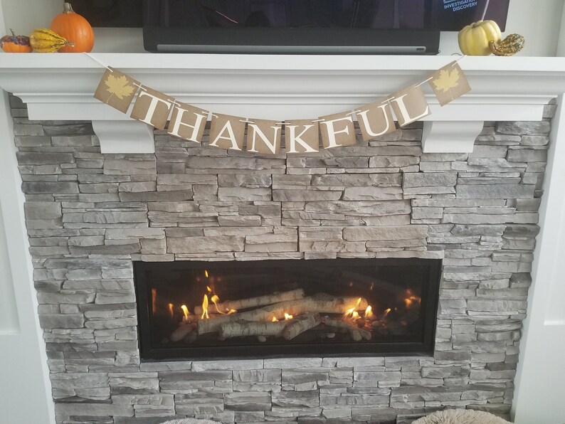 K&ouml;nnte beinhalten: Ein Kamin mit einem Steinsockel und einem Banner mit der Aufschrift "THANKFUL", das dar&uuml;ber h&auml;ngt. Der Kamin ist mit einem Feuer entz&uuml;ndet und es befinden sich Holzscheite im Kamin.