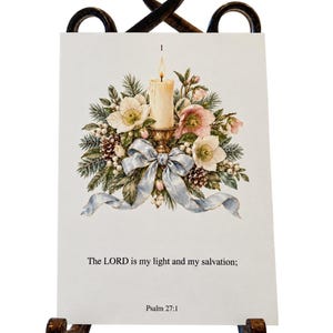 May include: A rectangular card on a dark metal stand featuring a lit candle, pine branches, white and pink flowers, pinecones, and a light blue ribbon. The text reads: 1, The LORD is my light and my salvation; Psalm 27:1.