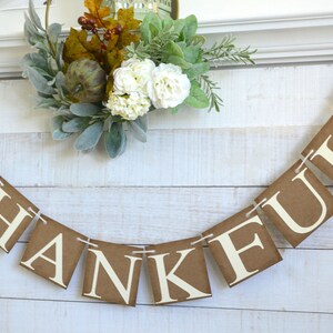 K&ouml;nnte beinhalten: Ein braunes Papierbanner mit dem Wort "THANKFUL" in wei&szlig;en Buchstaben. Das Banner ist mit wei&szlig;em Garn befestigt und hat an jedem Ende ein Herbstblatt-Design.