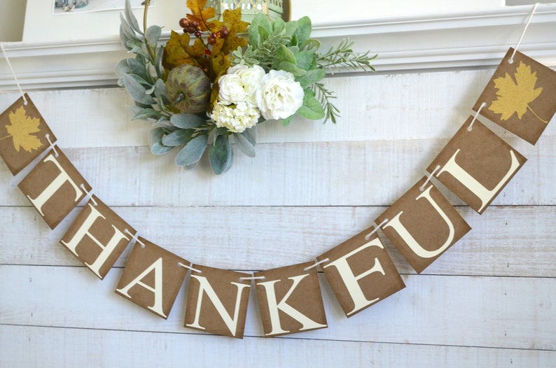 K&ouml;nnte beinhalten: Ein braunes Papierbanner mit dem Wort "THANKFUL" in wei&szlig;en Buchstaben. Das Banner ist mit wei&szlig;em Garn befestigt und hat an jedem Ende ein Herbstblatt-Design.