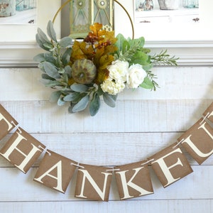 K&ouml;nnte beinhalten: Ein braunes Papierbanner mit dem Wort "THANKFUL" in wei&szlig;en Buchstaben. Das Banner ist mit Schnur befestigt und mit Herbstbl&auml;ttern verziert.