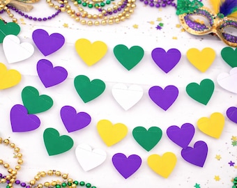 May include: A festive arrangement featuring heart-shaped decorations in green, purple, white, and yellow. The hearts are arranged in rows, complemented by beaded necklaces and a decorative mask, evoking a celebratory theme.
