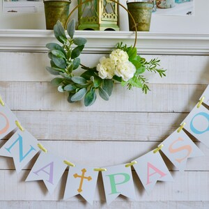 May include: A white wooden mantel with a yellow ribbon banner that reads "BUONA PASQUA" in pastel colors. The banner is decorated with a gold cross. A green and white floral wreath hangs above the banner.