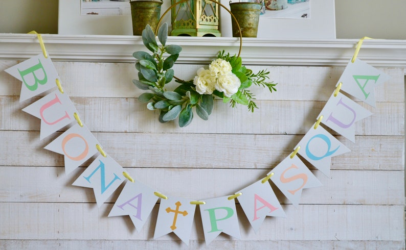 May include: A white wooden mantel with a yellow ribbon banner that reads "BUONA PASQUA" in pastel colors. The banner is decorated with a gold cross. A green and white floral wreath hangs above the banner.
