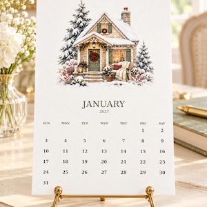May include: A January 2027 calendar page featuring a watercolor illustration of a snow-covered cottage with pine trees, a wreath on the door, and a cozy porch setting. The calendar grid displays the days of the month clearly in black text.