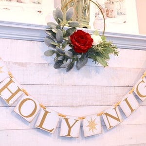 May include: A white wooden sign with gold lettering that reads "O Holy Night" with a gold star on each side of the word "Holy". The sign is hanging on a white wooden wall with a small green and red floral arrangement above it.