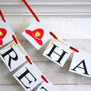 K&ouml;nnte beinhalten: Ein rot-wei&szlig;es Banner mit dem Wort "Happy" in schwarzen Buchstaben. Jeder Buchstabe befindet sich auf einem separaten quadratischen Papierst&uuml;ck mit einem goldenen Design in der Ecke. Das Banner ist mit einem roten Band zusammengef&uuml;gt.