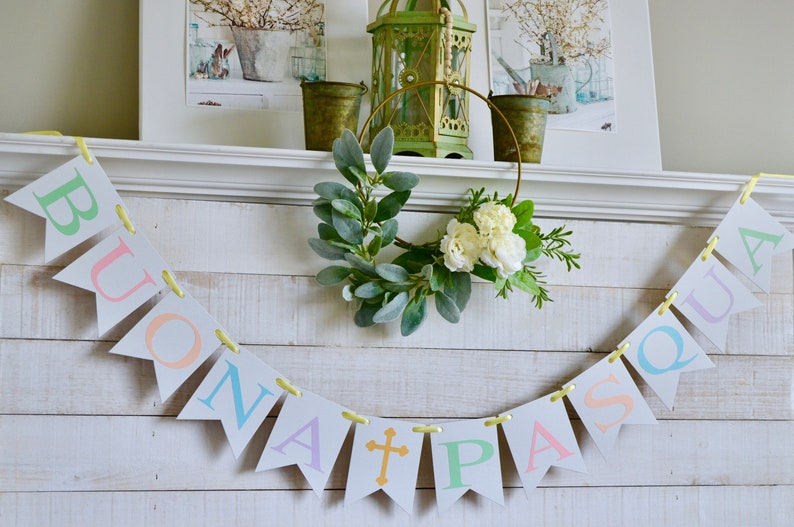 May include: A white wooden mantel with a banner that reads "BUONA PASQUA" in pastel colors. The banner is strung with yellow ribbon. A wreath with white flowers and green leaves hangs above the banner.