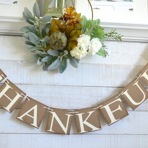 K&ouml;nnte beinhalten: Ein braunes Papierbanner mit dem Wort "THANKFUL" in wei&szlig;en Buchstaben. Das Banner ist mit goldenen Glitzerbl&auml;ttern verziert und mit wei&szlig;em Garn befestigt. Das Banner h&auml;ngt an einer wei&szlig;en Holzwand.