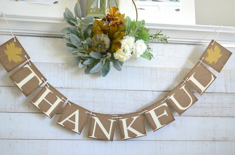 K&ouml;nnte beinhalten: Ein braunes Papierbanner mit dem Wort "THANKFUL" in wei&szlig;en Buchstaben. Das Banner ist mit goldenen Glitzerbl&auml;ttern verziert und mit wei&szlig;em Garn befestigt. Das Banner h&auml;ngt an einer wei&szlig;en Holzwand.