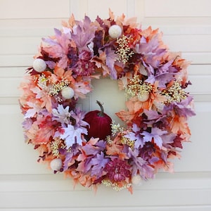 Peut inclure: Une couronne circulaire composée d'un arrangement dense de feuilles d'érable artificielles dans des tons orange, violet et rose clair. La couronne est agrémentée de petits éléments floraux sphériques blancs et d'une citrouille en velours rouge foncé.