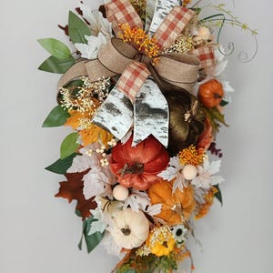 May include: A teardrop-shaped autumn swag featuring a mix of velvet pumpkins in orange, rust, and cream colours. The arrangement includes faux maple leaves, birch-patterned ribbon, hessian bows, and small sprigs of dried-style foliage and berries.