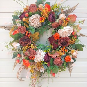 Peut inclure: Une couronne circulaire composée d'un arrangement dense de fleurs artificielles dans des tons crème, bordeaux profond et orange vif. Le design comprend divers feuillages verts, des brins de fougère bruns et des grappes de petites baies orange et rouges.