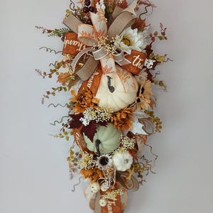 Peut inclure: Une guirlande verticale en forme de larme pour l'automne avec une grande citrouille crème, une citrouille vert sauge plus petite et une minuscule citrouille blanche. L'ensemble inclut des fleurs en soie orange et marron, du blé et un ruban en toile de jute.