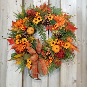 May include: A circular autumn wreath featuring orange and yellow flowers, small pumpkins, pinecones, and green fern foliage. A plaid ribbon in shades of orange, green, and brown is tied at the bottom, adding texture to the seasonal arrangement.