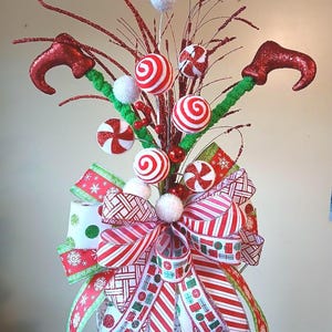 May include: A decorative festive topper featuring two red glittery elf legs, white and red peppermint-style spheres, green fuzzy stems, and a large multi-layered bow with red, white, and green patterns including snowflakes and gift boxes.