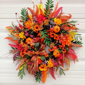 Peut inclure: Une couronne décorative composée d'un mélange vibrant de fleurs artificielles aux couleurs automnales, de feuillage vert, de pommes de pin, de petites citrouilles orange et de baies rouges. Un nœud en tissu écossais est centré en bas.