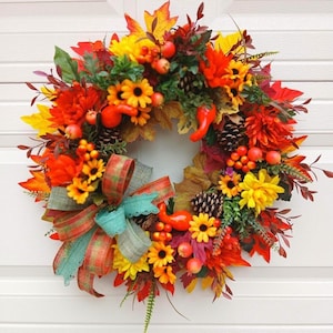 Peut inclure: Une couronne d'automne circulaire composée d'un mélange vibrant de tournesols jaunes et orange, de dahlias rouges, de pommes de pin brunes et d'un feuillage automnal varié. L'ensemble est agrémenté de petites baies orange et d'un nœud.