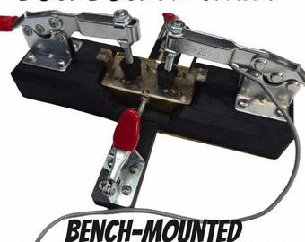 Könnte beinhalten: Eine schwarze, auf der Werkbank montierte Lötvorrichtung für Humbucker mit drei Metall-Kniehebelspannern mit roten Griffen. Das Gerät ist mit BUCKBUCKER CLAMP und BENCH-MOUNTED HUMBUCKER SOLDERING JIG beschriftet und hält einen metallischen Tonabnehmer.