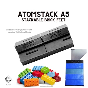 May include: Two black plastic Atomstack A5 stackable brick feet. The feet are designed to raise and lower a laser with standard 2x4 bricks. The feet have the text "KGD" printed on them. A pile of colourful plastic bricks are in the foreground. The text "Bricks not included" is printed below the pile of bricks.