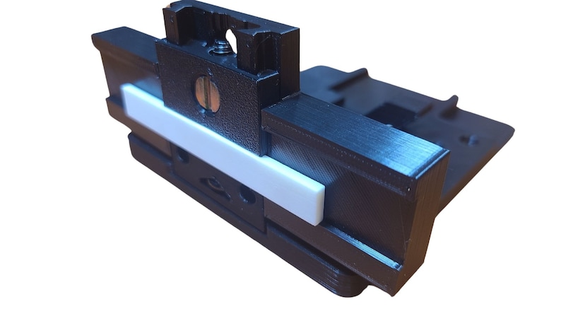 K&ouml;nnte beinhalten: Ein schwarzer 3D-gedruckter Kamerahalter mit einem wei&szlig;en Kunststoff-Schieber. Der Halter hat ein Schraubenloch und einen Schlitz f&uuml;r eine Kamera.