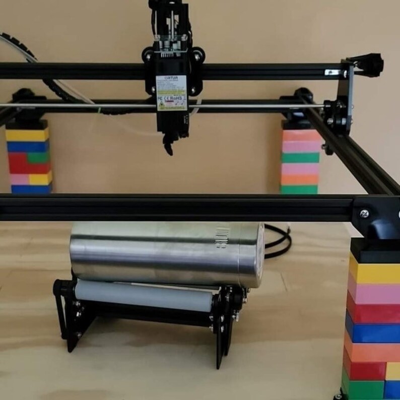 May include: A black laser cutter with a silver metal cylinder on a roller stand. The laser cutter is mounted on a black frame with colorful Lego blocks supporting the frame.