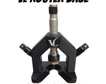 Könnte beinhalten: Eine schwarze VL-Fräserbasis mit einer silbernen Mittelsäule und zwei seitlichen Aufsätzen. Der Text VL ROUTER BASE und FITS SM FOREDOM-HANDPIECES ist in Schwarz aufgedruckt. Das Gerät verfügt über einen zentralen Einstellknopf mit VL-Logo.