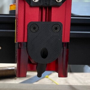 K&ouml;nnte beinhalten: Nahaufnahme eines roten und schwarzen Laserschneidkopfes. Der Kopf hat einen schwarzen Kunststoffschild mit zwei Schrauben und einer D&uuml;se. Der Laserschneider ist auf einer Holzoberfl&auml;che montiert.