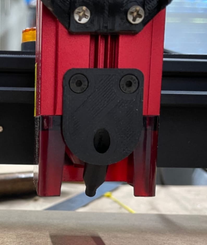 K&ouml;nnte beinhalten: Nahaufnahme eines roten und schwarzen Laserschneidkopfes. Der Kopf hat einen schwarzen Kunststoffschild mit zwei Schrauben und einer D&uuml;se. Der Laserschneider ist auf einer Holzoberfl&auml;che montiert.