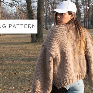 May include: A beige, chunky knit jumper with a wide neckline and long sleeves. The words "KNITTING PATTERN" are in a white rectangle. The jumper is worn by a person wearing a white baseball cap.